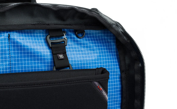 A close shot of the rear main pocket of a Black 525 Ballistic exterior Western Flyer opened to reveal an Island (blue) 200 Halcyon interior with a Cache connected to the nylon loops sewn into the interior seam and a nearby o-ring.